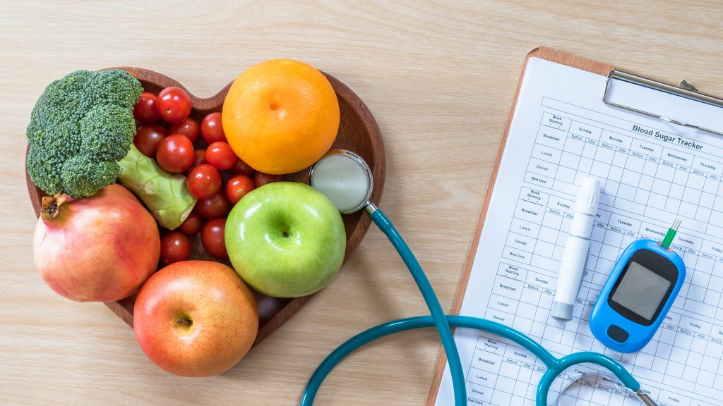 A heart shaped wooden bowl of fruit and vegetables beside a Blood Sugar tracker clipboard, stethoscope and a blood sugar monitoring device to represent Type 2 Diabetes